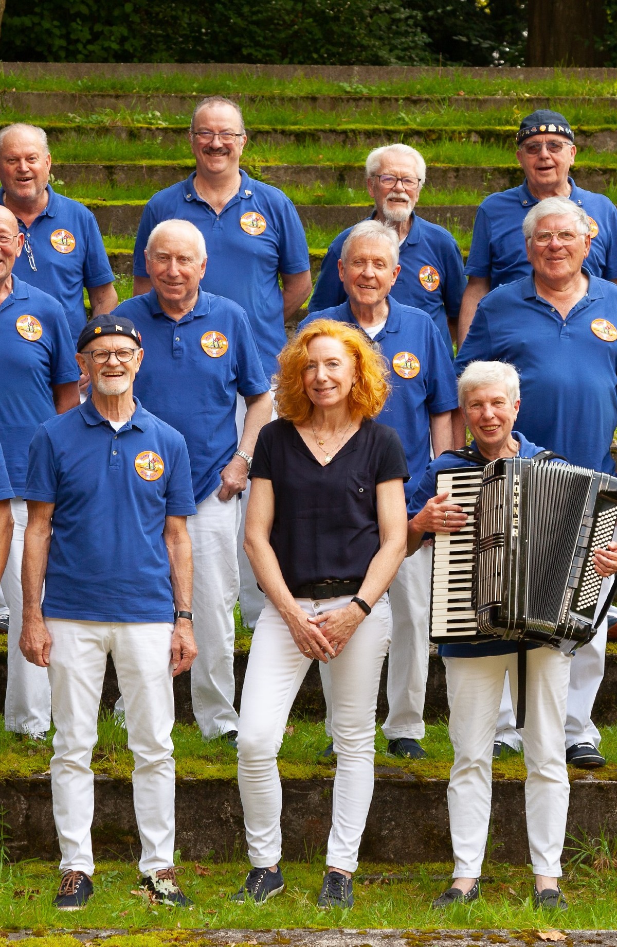 Der Shanty Chor in blauen Poloshirts und weißen Hosen posiert fröhlich auf Stufen im Freien; eine Frau und eine Frau mit einem Akkordeon stehen in der Mitte.