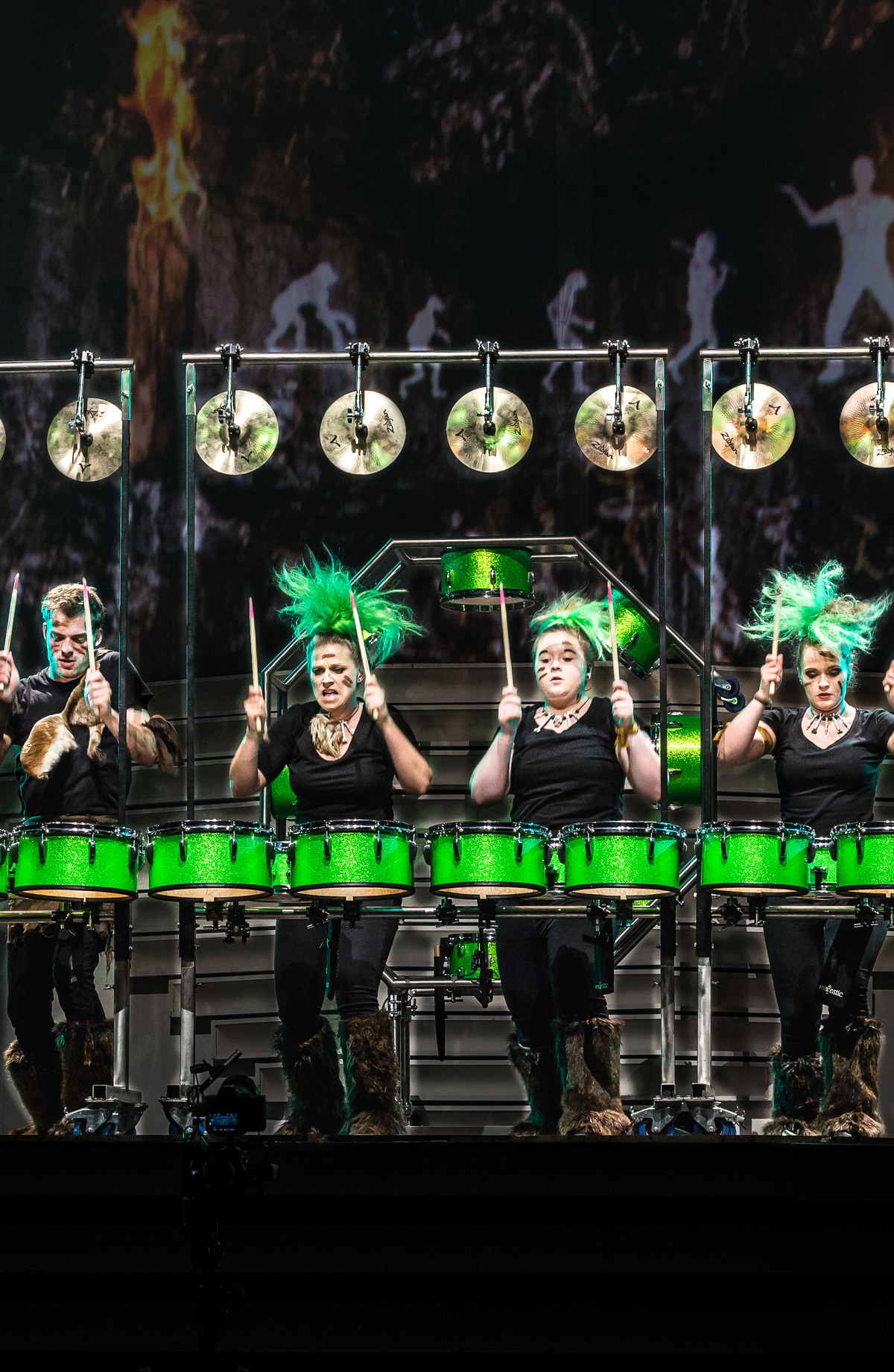 Sechs Trommler performen an einem beleuchteten Schlagzeug mit grünen Trommeln vor einem Hintergrund, der eine waldähnliche Szene zeigt. Sie sind energisch und tragen einheitliche schwarze Outfits mit grünen Akzenten.
