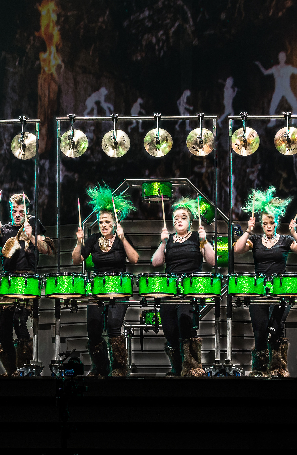 Sechs Trommler performen an einem beleuchteten Schlagzeug mit grünen Trommeln vor einem Hintergrund, der eine waldähnliche Szene zeigt. Sie sind energisch und tragen einheitliche schwarze Outfits mit grünen Akzenten.