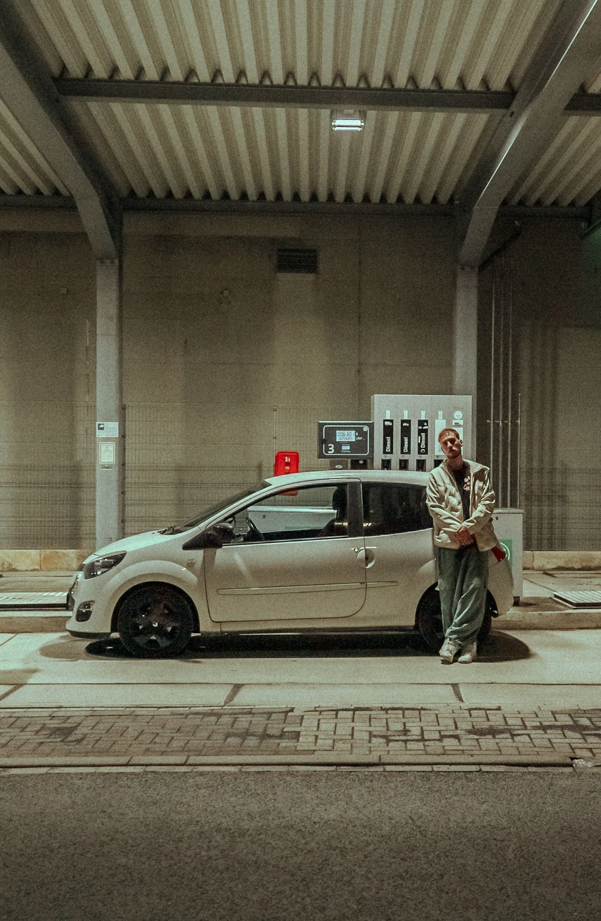 Sebastian Gröne aka Tänzer hockt vor einem weißen Kleinwagen, der neben einer Tanksäue steht, in einer nächtlichen, urbanen Umgebung.