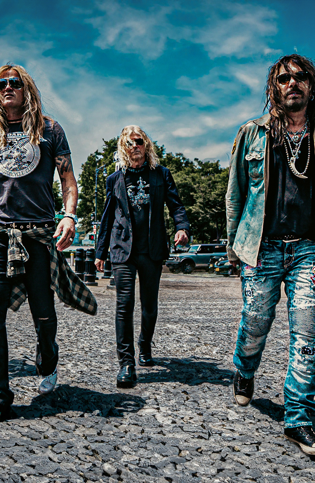 Ein Gruppe von fünf Personen, die wie eine Rockband gekleidet sind, geht auf einer Kopfsteinpflasterstraße mit einem bewölkten Himmel im Hintergrund.