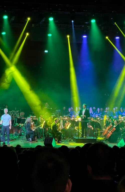 Ein Live-Konzert mit einer Band im Vordergrund und einem klassischen Orchester im Hintergrund, beleuchtet von dramatischen grünen und gelben Lichtstrahlen.