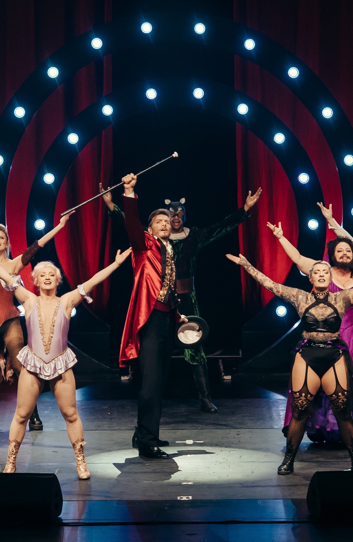 Darsteller in Kostümen auf der Bühne mit erhobenen Armen im Hintergrund leuchten Scheinwerfer vor einem roten Vorhang