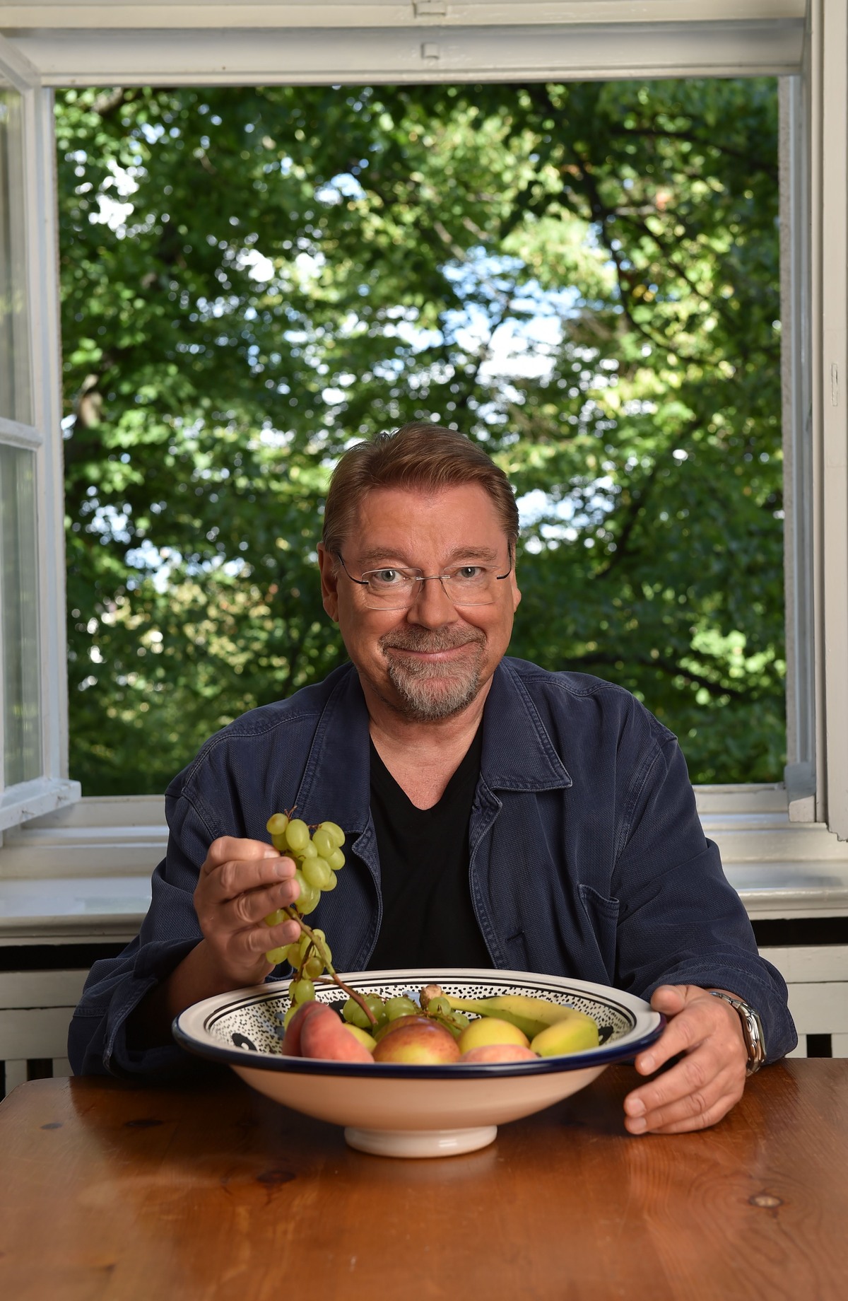 Jürgen von der Lippe am Tisch sitzend vor dem geöffneten Fenster. Vor ihm steht eine Schale mit Obst. Er hält Trauben in der Hand.