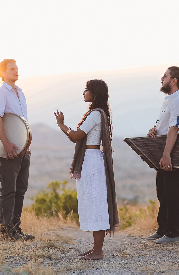 Die fünf Bandmitglieder, in weißer und brauner Kleidung, stehen seitlich nebeneinander mit Musikinstrumenten vor einem Sonnenuntergang.