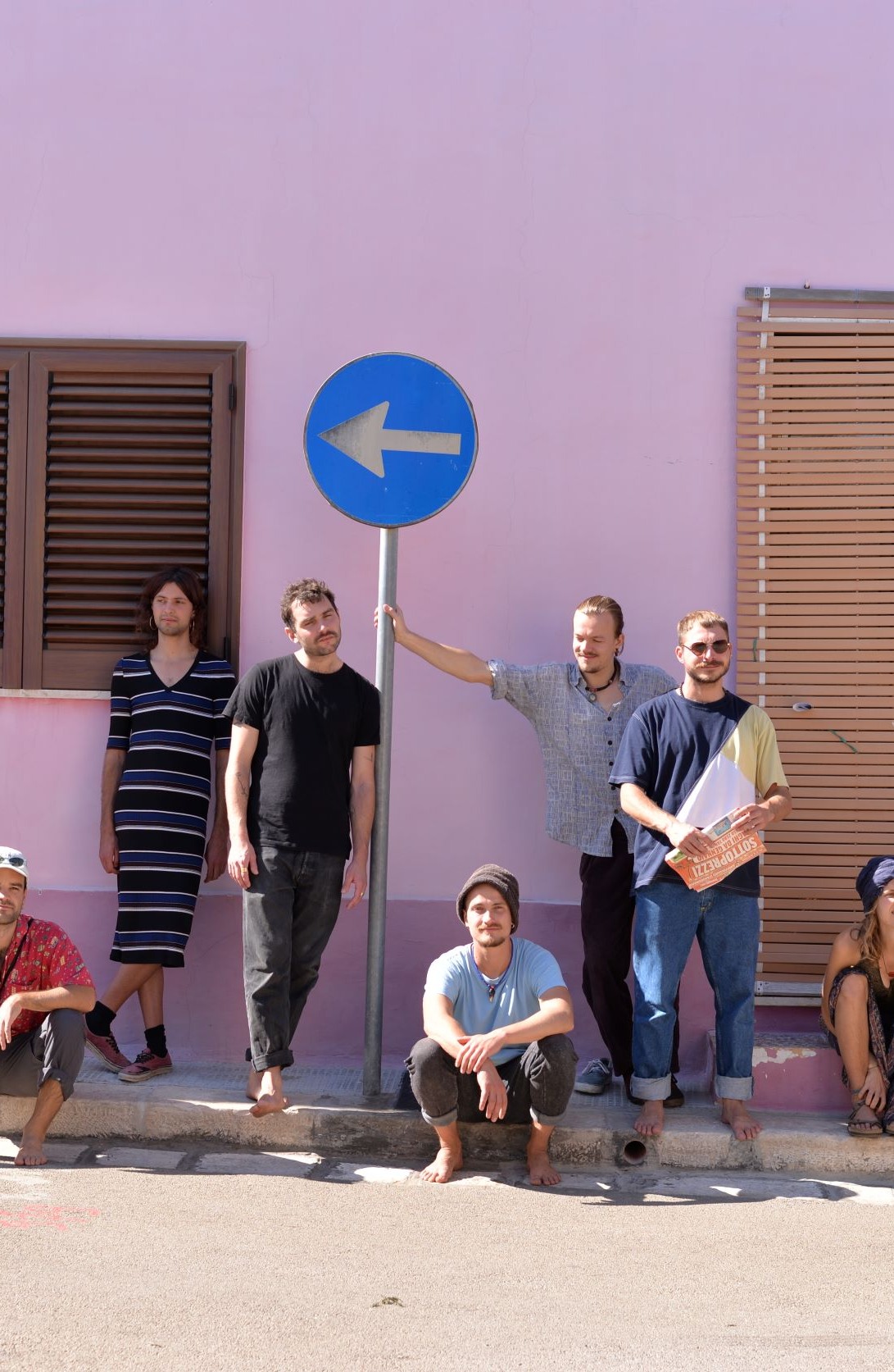 Bandmitglieder sitzen auf Bürgersteig vor einer rosa Hauswand