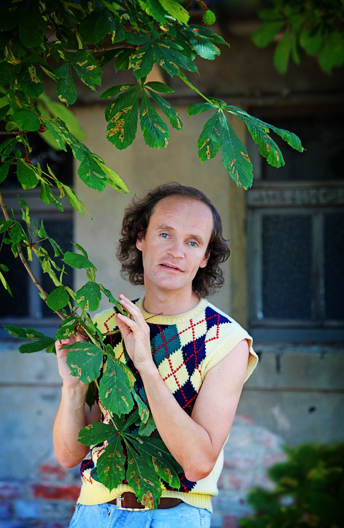 Olaf Schubert  in einem ärmellosen Pullover mit Argyle-Muster steht vor einem grünen Blätterhintergrund und hält vorsichtig einen Zweig. Er lächelt leicht und schaut direkt in die Kamera.