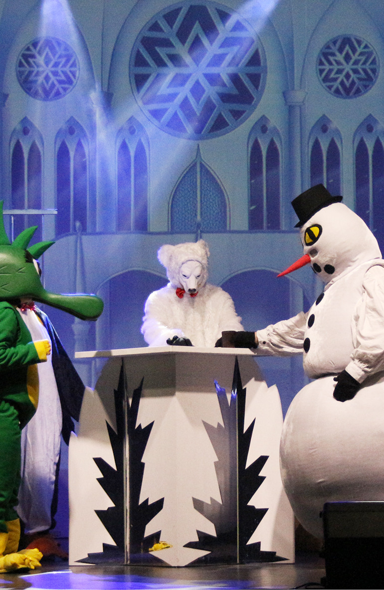 Personen in Kostümen auf einer Bühne mit märchenhaftem Hintergrund: ein grüner Drache, ein weißer Bär, ein Schneemann mit Zylinder und ein Pinguin, in einer Szene aus einem Theaterstück.