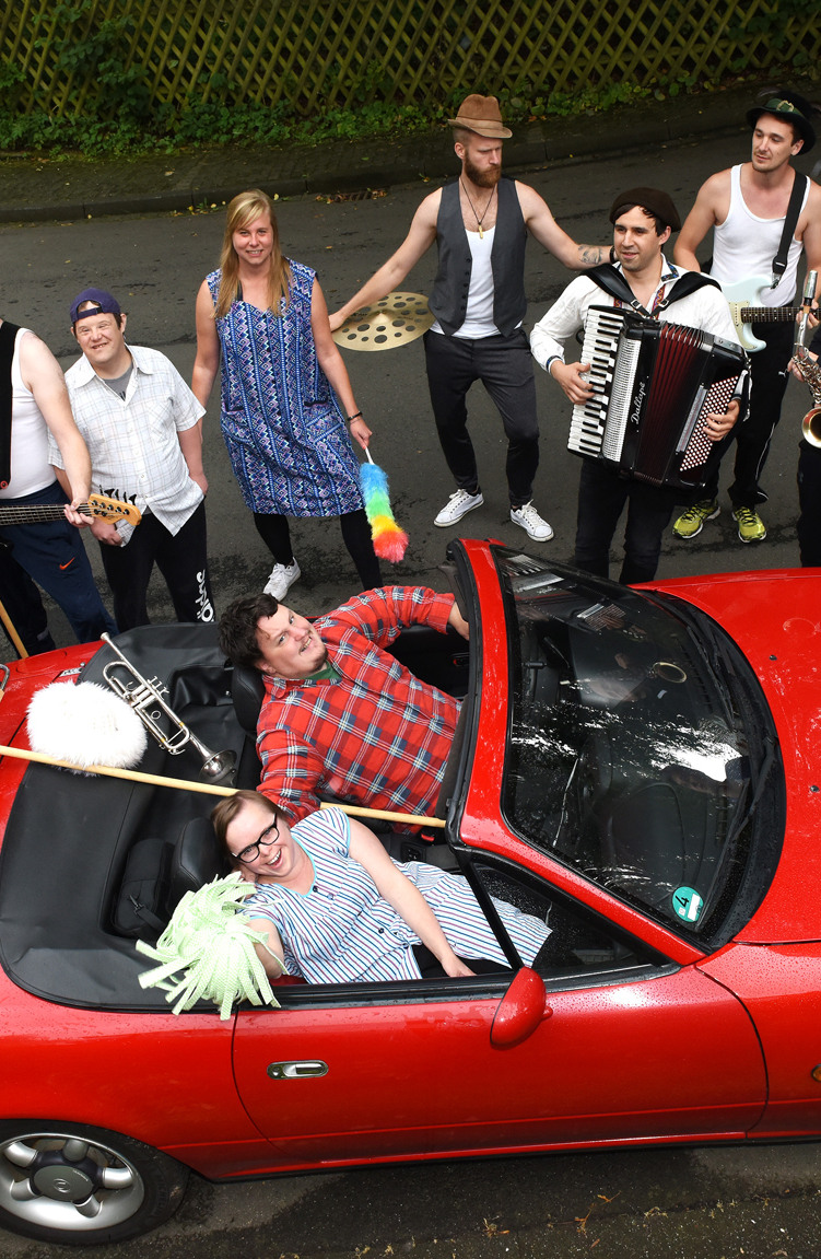 Eine fröhliche Gruppe von Menschen in verschiedenen Kostümen steht um ein rotes Cabrio herum, einige mit Musikinstrumenten.